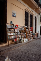 A narrow cobblestone street is lined with wooden racks displaying a variety of books and magazines. The racks are positioned against a light beige wall, with barred windows above them. A sign and posters add character to the scene. A small portion of a dog can be seen in the bottom left corner, enhancing the street market atmosphere.