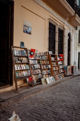 A narrow cobblestone street is lined with wooden racks displaying a variety of books and magazines. The racks are positioned against a light beige wall, with barred windows above them. A sign and posters add character to the scene. A small portion of a dog can be seen in the bottom left corner, enhancing the street market atmosphere.