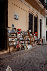 A narrow cobblestone street is lined with wooden racks displaying a variety of books and magazines. The racks are positioned against a light beige wall, with barred windows above them. A sign and posters add character to the scene. A small portion of a dog can be seen in the bottom left corner, enhancing the street market atmosphere.