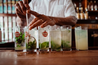 A bartender stirs a series of cocktails in tall glasses, each adorned with mint leaves and labeled with the Havana Club logo. The setting appears to be a dimly lit bar with bottles and a wooden counter.