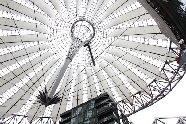 Looking up from below, a large, intricately designed roof structure is visible, featuring a radial pattern of white stretched fabric panels supported by a network of steel cables and beams. In the center, there's a circular opening with additional structural elements converging towards it. Below, multi-story modern buildings with glass facades are partially seen.