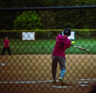 A baseball player in a pink jersey with the number 9 swings a bat at a softball. The action takes place behind a chain-link fence on a playing field with a grassy background. Another player is visible in the outfield.