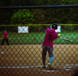 A baseball player in a pink jersey with the number 9 swings a bat at a softball. The action takes place behind a chain-link fence on a playing field with a grassy background. Another player is visible in the outfield.