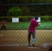 A baseball player in a pink jersey with the number 9 swings a bat at a softball. The action takes place behind a chain-link fence on a playing field with a grassy background. Another player is visible in the outfield.