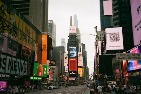 A bustling urban scene with numerous digital billboards and advertisements lining the buildings. The street is filled with walking pedestrians and some vehicles. Tall skyscrapers rise in the background, contributing to the vibrant city atmosphere. Prominent advertisements for products like Samsung and Coca-Cola are visible.
