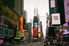 A bustling urban scene with numerous digital billboards and advertisements lining the buildings. The street is filled with walking pedestrians and some vehicles. Tall skyscrapers rise in the background, contributing to the vibrant city atmosphere. Prominent advertisements for products like Samsung and Coca-Cola are visible.
