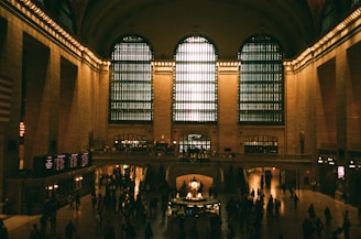 A large, dimly lit hall with high ceilings and expansive windows letting in natural light. People are scattered throughout, some walking, others standing near informational displays. A grand clock is positioned centrally, surrounded by warm yellow lighting.