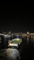 A ferry is traveling on a dark body of water at night. The city skyline is illuminated in the background, featuring prominent landmarks with their lights reflecting off the water's surface. The ferry has a well-lit deck and is making a gentle wake as it moves.