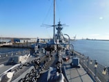 A large naval battleship is docked near an industrial area on a clear day. The ship's deck includes large chains, netting, and various equipment. In the background, there is a view of warehouses, a bridge, and cranes near the waterfront.