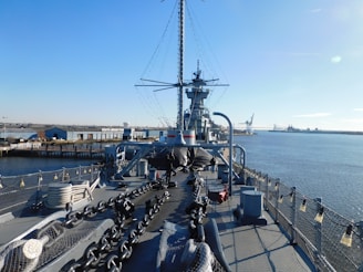 A large naval battleship is docked near an industrial area on a clear day. The ship's deck includes large chains, netting, and various equipment. In the background, there is a view of warehouses, a bridge, and cranes near the waterfront.