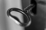 A close-up, black and white photograph of a key inserted into a lock. The focus is on the key's circular handle, highlighting its metallic texture and details.