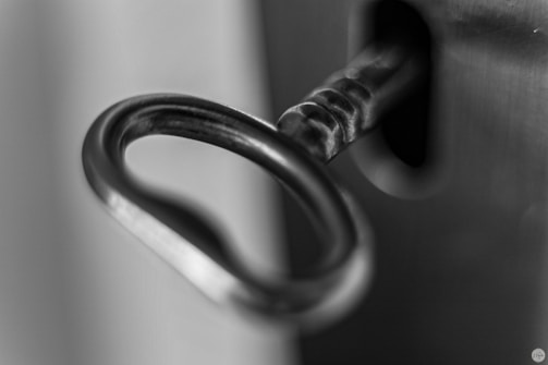 A close-up, black and white photograph of a key inserted into a lock. The focus is on the key's circular handle, highlighting its metallic texture and details.