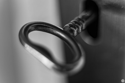 A close-up, black and white photograph of a key inserted into a lock. The focus is on the key's circular handle, highlighting its metallic texture and details.