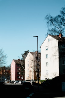 A row of multi-story residential buildings along a street with several parked cars. The buildings have a simple, geometric design with predominantly white and earthy-colored walls. Leafless trees stand in front of the buildings, and a clear blue sky provides a bright backdrop. The scene appears quiet and suburban.