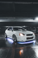 A sleek white car parked in a dimly lit garage. The car is equipped with custom rims and has blue neon lights underneath. The lighting highlights the glossy finish of the car. Overhead beams and reflective tiled flooring add to the industrial ambiance.
