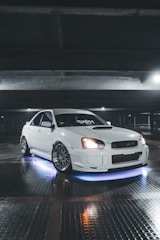 A sleek white car parked in a dimly lit garage. The car is equipped with custom rims and has blue neon lights underneath. The lighting highlights the glossy finish of the car. Overhead beams and reflective tiled flooring add to the industrial ambiance.