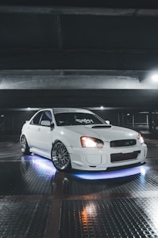 A sleek white car parked in a dimly lit garage. The car is equipped with custom rims and has blue neon lights underneath. The lighting highlights the glossy finish of the car. Overhead beams and reflective tiled flooring add to the industrial ambiance.