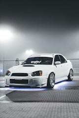 A sleek white car with custom modifications is parked under a foggy, illuminated environment. The car features distinctive headlights and rims, and there is an underglow of blue light creating a dramatic effect.
