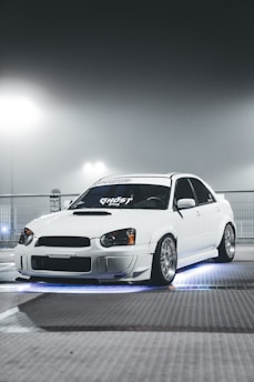 A sleek white car with custom modifications is parked under a foggy, illuminated environment. The car features distinctive headlights and rims, and there is an underglow of blue light creating a dramatic effect.