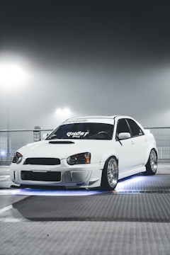 A sleek white car with custom modifications is parked under a foggy, illuminated environment. The car features distinctive headlights and rims, and there is an underglow of blue light creating a dramatic effect.