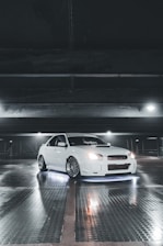 A white sports car with customized features is parked inside a dimly lit garage. The car is illuminated by underglow lights, creating a striking effect against the reflective, wet surface of the floor. Overhead lighting casts subtle shadows and highlights the sleek design of the vehicle.