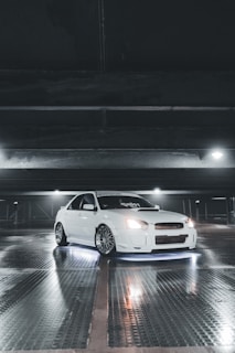 A white sports car with customized features is parked inside a dimly lit garage. The car is illuminated by underglow lights, creating a striking effect against the reflective, wet surface of the floor. Overhead lighting casts subtle shadows and highlights the sleek design of the vehicle.