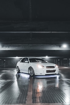 A white sports car with customized features is parked inside a dimly lit garage. The car is illuminated by underglow lights, creating a striking effect against the reflective, wet surface of the floor. Overhead lighting casts subtle shadows and highlights the sleek design of the vehicle.