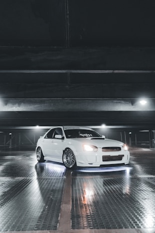 A white sports car with customized features is parked inside a dimly lit garage. The car is illuminated by underglow lights, creating a striking effect against the reflective, wet surface of the floor. Overhead lighting casts subtle shadows and highlights the sleek design of the vehicle.