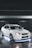 A white sports car with a modified body kit is parked on a reflective surface. It is equipped with large alloy wheels and features underglow lighting that casts a blue hue on the ground. The background is a dimly lit parking structure.