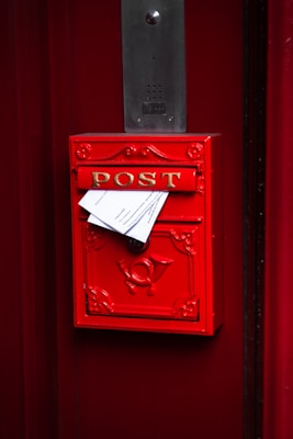 A bright red mailbox with the word 'POST' in gold letters is mounted on a red door. Papers are protruding from the mail slot, and a decorative emblem is visible on the front. Above the mailbox, there is a rectangular metal intercom panel.