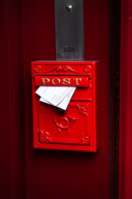 A bright red mailbox with the word 'POST' in gold letters is mounted on a red door. Papers are protruding from the mail slot, and a decorative emblem is visible on the front. Above the mailbox, there is a rectangular metal intercom panel.
