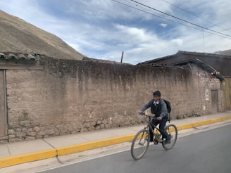 A young boy is riding a bicycle along a paved road. He is wearing a gray sweater, formal pants, and has a backpack. The background features an old rustic wall and a building with a tin roof. The sky is partly cloudy, and the scene appears to be rural.