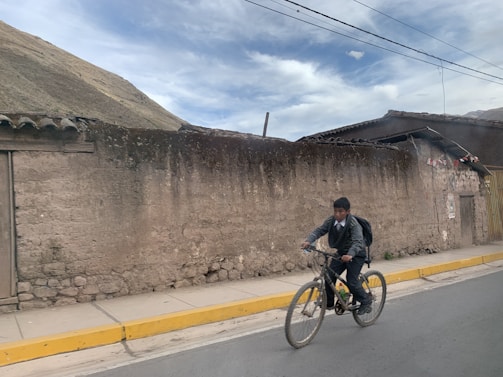 A young boy is riding a bicycle along a paved road. He is wearing a gray sweater, formal pants, and has a backpack. The background features an old rustic wall and a building with a tin roof. The sky is partly cloudy, and the scene appears to be rural.