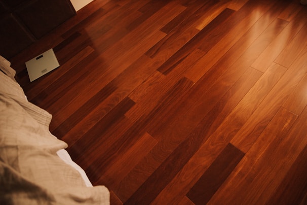A view of a polished wooden floor with rich, dark brown hues. To the left, there is a corner of a bed with light-colored sheets, and a digital scale is positioned near the bed on the floor.