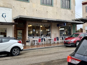 A small diner with a rustic appearance has a sign advertising fried chicken and fried green tomatoes. Outdoor seating is available with metal chairs and tables. Several vehicles are parked outside on a wet street, indicating recent rain. The building has a vintage facade with green window frames and a sign for 'Pearl's'.
