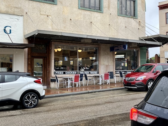 A small diner with a rustic appearance has a sign advertising fried chicken and fried green tomatoes. Outdoor seating is available with metal chairs and tables. Several vehicles are parked outside on a wet street, indicating recent rain. The building has a vintage facade with green window frames and a sign for 'Pearl's'.