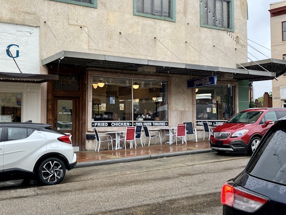 A small diner with a rustic appearance has a sign advertising fried chicken and fried green tomatoes. Outdoor seating is available with metal chairs and tables. Several vehicles are parked outside on a wet street, indicating recent rain. The building has a vintage facade with green window frames and a sign for 'Pearl's'.