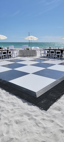 A beachside setup featuring a large black and white checkered dance floor on sand, surrounded by white chairs and tables with umbrellas offering shade. The ocean is visible in the background under a clear blue sky.