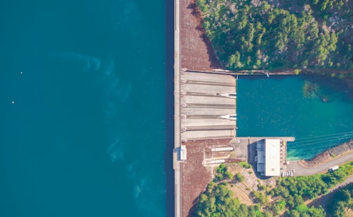 An aerial view of a large dam situated between a vast body of water and a lush forest. The structure of the dam includes spillways and a building at one end. The forested area is dense with green trees.