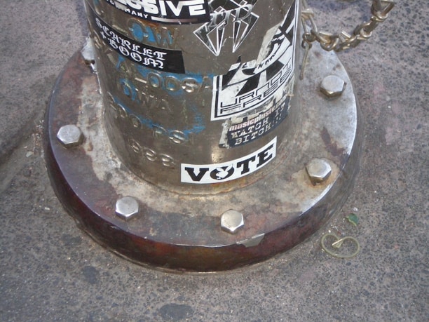 A metal pole base with large bolts and various stickers attached. The surface is weathered, featuring a prominent sticker that reads 'VOTE' in bold letters. Other stickers display graffiti-style text and graphics.