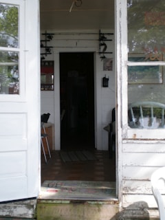 A rustic interior space featuring a view through an open door into a dimly lit hallway. On the walls, fishing rods are mounted horizontally. There's a white wooden door and window frame, with a worn-out appearance. A black chair is seen inside, and a mat with faded colors lies at the doorstep.