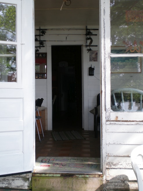 A rustic interior space featuring a view through an open door into a dimly lit hallway. On the walls, fishing rods are mounted horizontally. There's a white wooden door and window frame, with a worn-out appearance. A black chair is seen inside, and a mat with faded colors lies at the doorstep.