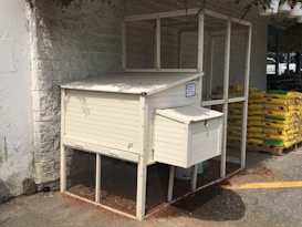 A white chicken coop is situated against a light-colored brick wall. The coop is enclosed with a mesh structure and features a small lockable compartment on the side. In the background, there are several bags of potting mix stacked on pallets. Some hanging plants are partially visible above, casting shadows on the coop.
