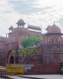 The image features an ancient red sandstone fort with intricate architectural details, including domes and arched gateways. The fort is surrounded by lush green foliage and a clear sky with scattered clouds. A yellow barrier marked 'Delhi Police' is visible in the foreground, along with a tree bearing flowers.