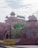 The image features an ancient red sandstone fort with intricate architectural details, including domes and arched gateways. The fort is surrounded by lush green foliage and a clear sky with scattered clouds. A yellow barrier marked 'Delhi Police' is visible in the foreground, along with a tree bearing flowers.