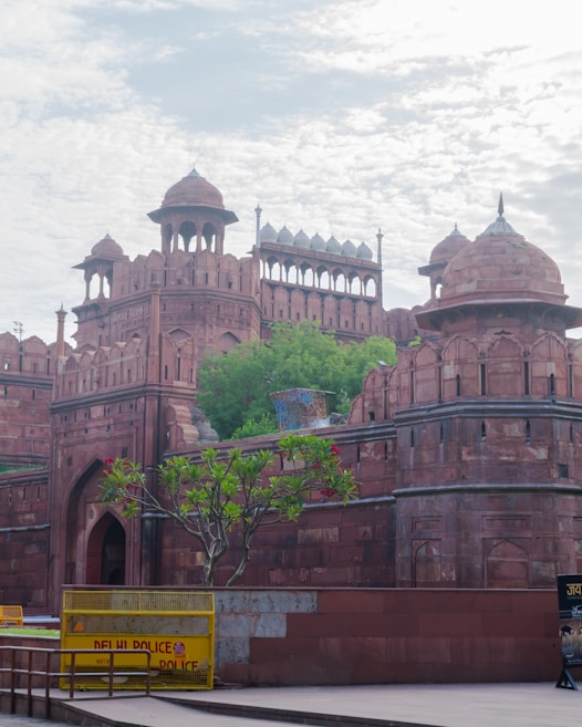 The image features an ancient red sandstone fort with intricate architectural details, including domes and arched gateways. The fort is surrounded by lush green foliage and a clear sky with scattered clouds. A yellow barrier marked 'Delhi Police' is visible in the foreground, along with a tree bearing flowers.