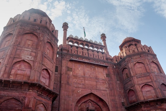 A grand red sandstone fort with large arched entrances and intricate architectural details. The fort has domed towers and balconies, with a partial view of a cloudy sky in the background.