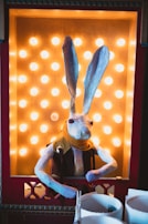 A papier-mâché rabbit figure with tall ears and red eyes is positioned inside a brightly lit display with golden lightbulbs. The rabbit is dressed in a brown vest and a yellow scarf, and it leans slightly forward. The display is framed by a red border with decorative elements.