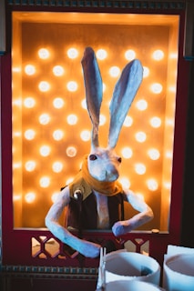 A papier-mâché rabbit figure with tall ears and red eyes is positioned inside a brightly lit display with golden lightbulbs. The rabbit is dressed in a brown vest and a yellow scarf, and it leans slightly forward. The display is framed by a red border with decorative elements.