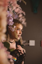 A person with blonde hair and facial piercings stands amidst a backdrop of pink and purple flowers. The lighting casts warm tones, and the person wears a lacy black outfit with a choker. A subtle rainbow detail is visible on their sleeve.